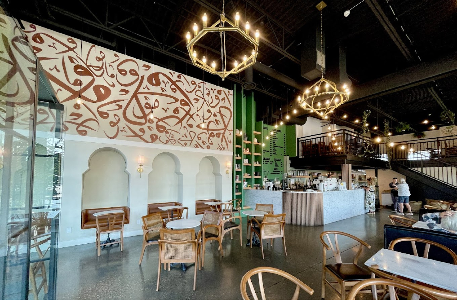 Interior of Mokha Coffee House, showing plenty of neutral toned seating, brown and white Arabic script bordering the ceiling, and seating alcoves with arches. Intérieur du café Mokha, avec de nombreux sièges aux tons neutres, des écritures en arabe brunes et blanches bordant le plafond, et des alcôves avec des arches.