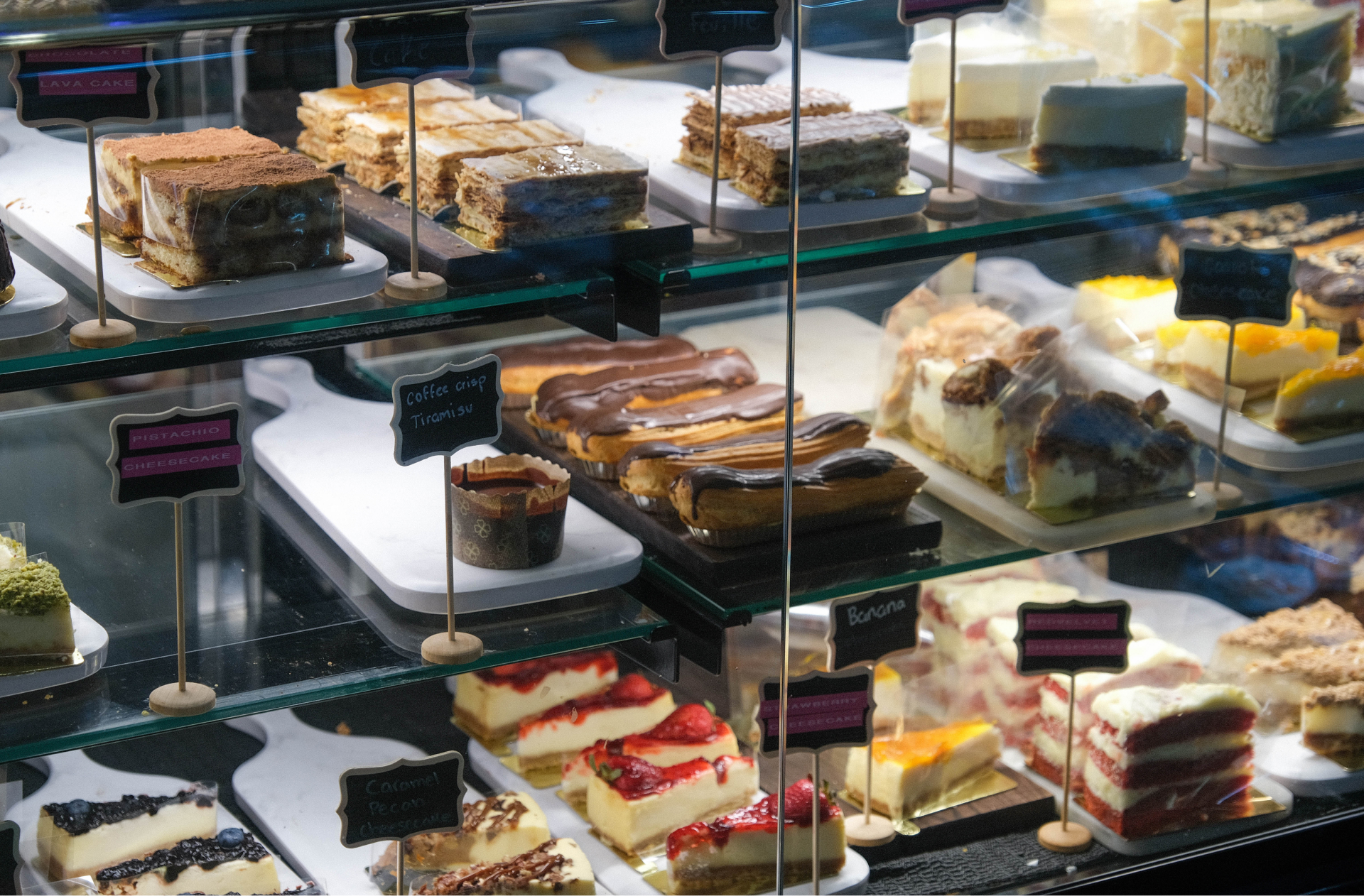 A close up of the dessert display case, with rows of cheesecakes, cakes, eclairs, and tiramisu. Gros plan sur le présentoir des desserts, avec des rangées de gâteau au fromage, de gâteaux, d’éclairs et de tiramisu.